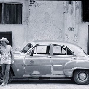Cuban man leaning against car smoking cigar (B&W)