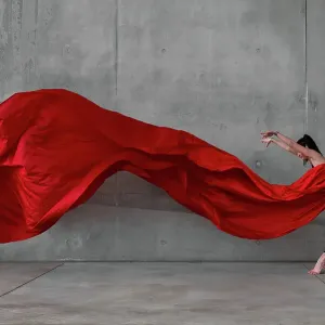Female ballet dancer on pointes dancing with a large red veil in front of a grey concrete wall