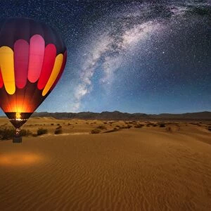 A majestic hot air balloon soars under the stars of the Milky Way, over the desert - Mesquite Dunes of Death Valley National Park. Moonlight provides luminosity showing the patterns and shapes of the desert landscape