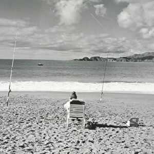 Person sitting on beach with fishing poles embedded in sand and using cell phone
