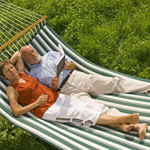 Senior citizen couple lying in a hammock, woman listening to music, man reading a book