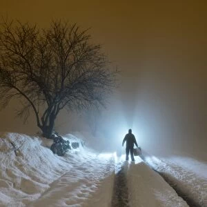 Silhouette of a person walking with a suitcase along a covered way of snow and fog during the night