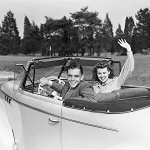 Teenage couple in convertible car smile and wave to viewer