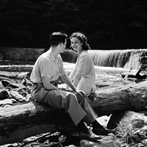 Teenage couple sitting on fallen log by side of creek, talking