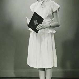 Teenage girl (13-14) in white dress standing in studio with open book, (B&W)
