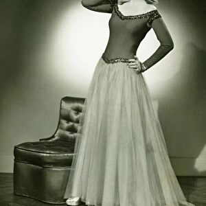 Woman in evening dress standing indoors, holding hand on cheek, (B&W)