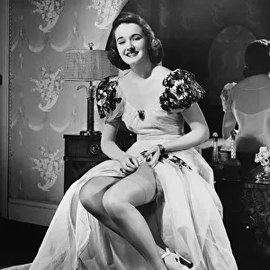 Woman in evening gown putting on stockings at vanity table (B&W), portrait
