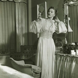 Woman in fashionable gown looking at mirror in bedroom, (B&W)