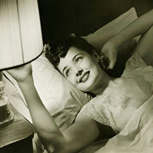 Woman lying on bed, turning off lamp on night table, (B&W), elevated view