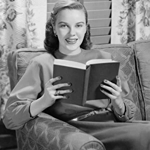 Woman reading book in living room, (B&W), portrait