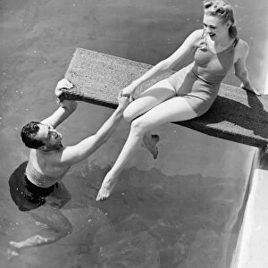Woman sitting on diving board, man grasping her hand (B&W), elevated view