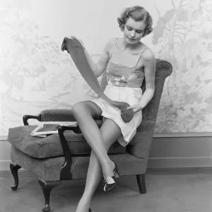 Woman wearing lingerie, sitting on arm of chair, holding up pair of silk stockings