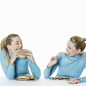 Two women eating food, one eating fast food, the other one eating uncooked vegetarian food