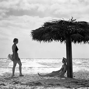 Two women under palm thatch umbrella on beach, Florida