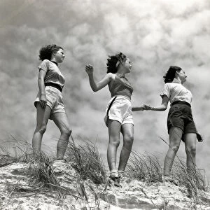 Three women standing on beach, holding hands, smiling. (Photo by H