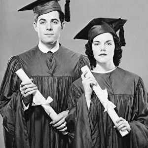 Young couple in graduate dress holding diplomas, posing in studio, (B&W), portrait