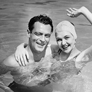 Young couple standing in pool, woman waving hand, (B&W), portrait