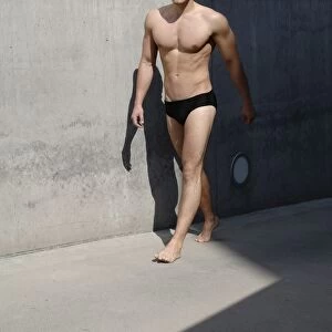 Young man wearing bathing trunks, walking in front of a concrete wall