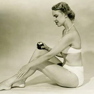 Young woman in bikini sitting, putting cream on leg, (B&W)