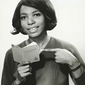 Young woman pointing at bank book, smiling, (B&W), portrait