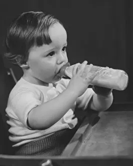 Baby girl (9-12 months) sitting in highchair, drinking milk from bottle (B&W)