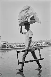 Prints of A barefoot man carrying builders trestles in one hand, using his other hand to support a load carried on a platform on his head, walking on wet ground in Saigon, Vietnam, 30th August 1965 Vietnam Prints: Barefoot In Saignon