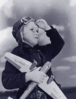 Boy (8-10) wearing flying cap and goggles holding toy plane (B&W)