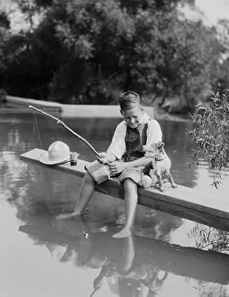 Non Urban Scene Gallery: Boy fishing with homemade pole, feet dangling in water, dog sitting by his side