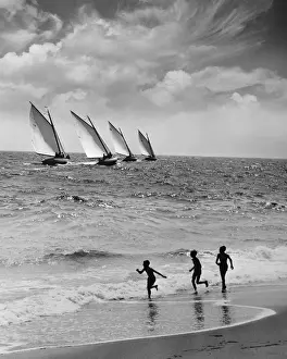 Three boys running along beach, following four sailboats out on ocean