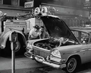 Prints of A fireman wearing a white t-shirt and a helmet leaning over the engine compartment of a Dodge car, with the hood raised to reveal a fire, a fire engine in the background, in Williamsport Timeless Moments: A Visual Journey Through History’s Defining Images: Checking Under The Hood