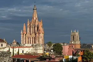 San Miguel De Allende Prints: Church and cityscape under cloudy sky, with a hot air balloon in the distance