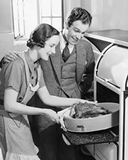 Couple in kitchen, wife taking roast from oven (B&W)