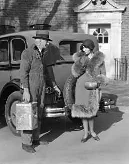 Couple. The Man Is Wearing A Fedora & Coveralls With 1 Hand On The Gas Tank Lid Of A Sedan & The Other Holding A Can The Woman A Cloche & A Fur Trim Coat