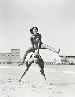 Retrofile Gallery: Couple playing leapfrog on beach, woman jumping over man