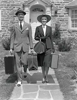 Couple walking along sidewalk, carrying suitcases