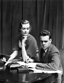 Serious Prints: Couple Working On Budget At Desk Woman Sitting On Arm Of Chair Man With Pencil Writing On