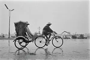 Prints of A cycle rickshaw riding through the rain, the driver wearing a safari-style hat, on an otherwise deserted street in Saigon, Vietnam, 30th August 1965 Vietnam Prints: Cycle Rickshaw, Saignon