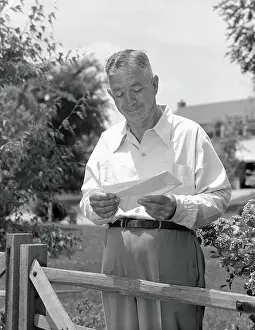 Mail Prints: Elderly man standing by fence in yard, opening envelope and taking out cheque