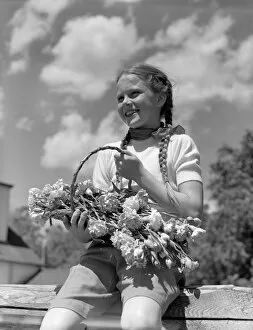 Recreational Pursuit Collection: Farm girl with pigtails, sitting on fence, holding basket of flowers, smiling