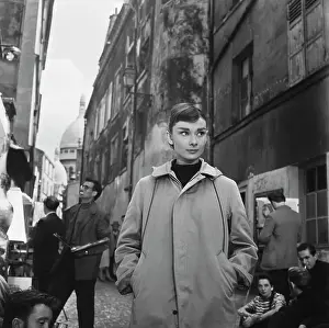 Prints of British actress Audrey Hepburn (1929-1993), her hands in the pockets of her raincoat, on a busy street, the dome of Sacre-Coeur visible in the background, filming Funny Face in Paris, France Elegance Personified – A Classic Art Print Collection: Filming Funny Face, 1956