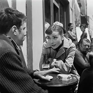 Prints of British actress Audrey Hepburn, wearing a raincoat, her elbows resting on the table before her as she sits outside a bar with a man during the filming of Funny Face in Paris, France, July 1956 Elegance Personified – A Classic Art Print Collection: Filming Funny Face, 1956