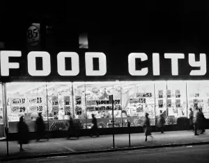 Prints of Pedestrians on the sidewalk pass the illuminated windows of a Food City store, with offers advertised in the store windows, United States, circa 1955 Chasing Speed: Motorsport in Motion: Food City