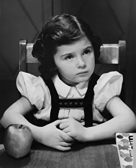 Girl (6-7) sitting at table, looking up, (B&W)