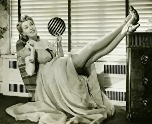 Hand Prints: Glamorous woman in evening gown holding hand mirror, legs on cabinet, (B&W), portrait