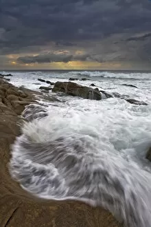 Landscape photo of ocean waves crashing onto the rocks under a stormy moody sky at sunrise