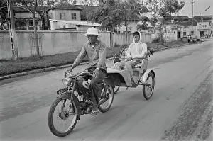 Prints of A motorcycle rickshaw driver, wearing a safari-style hat, driving a passenger through the street of Saigon, Vietnam, 30th August 1965 Vietnam Prints: Life In Saigon