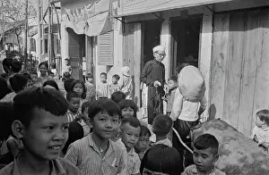 Prints of Vietnamese children on a rundown street, to the right of the image is a man wearing a black ao dai and holding a closed umbrella by a doorway alongside someone wearing a traditional non la hat Vietnam Prints: Life In Saigon