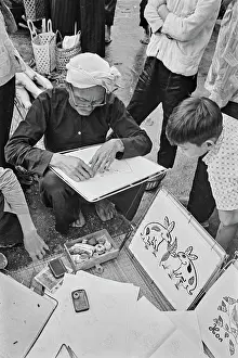 Prints of A child watches as an elderly Vietnamese man, wearing a white turban, draws using a drawing board, at his side is a picture of rabbits, on a street in Saigon, Vietnam, 30th August 1965 Vietnam Prints: Life In Saigon