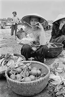 Prints of A Vietnamese woman, wearing a traditional non la hat, holds her baby in her arm as she sells vegetable from a basket on an embankment of the Saigon River as it flows through Saigon, Vietnam Vietnam Prints: Life Along The Saigon River