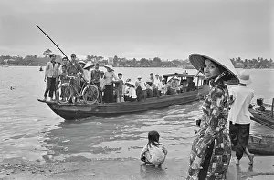 Prints of A Vietnamese woman, wearing a traditional non la hat, stands on the riverbank as a sampan ferrying passengers across the Saigon River moors in Saigon, Vietnam, 30th August 1965 Vietnam Prints: Life Along The Saigon River